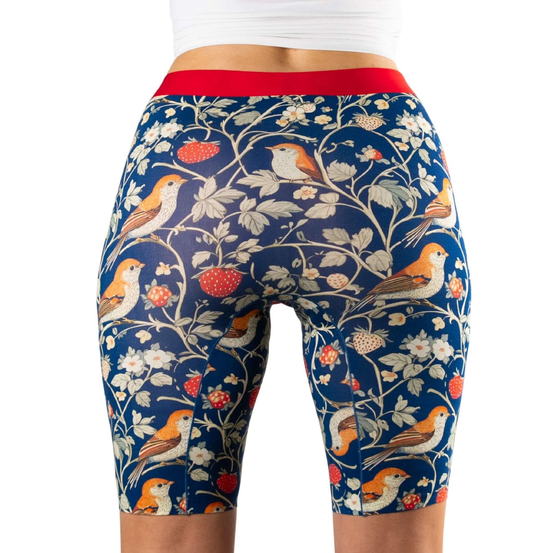 A woman wearing high-waisted biker shorts with a strawberries and bird print, featuring a vibrant red waistband and a dark blue background.
