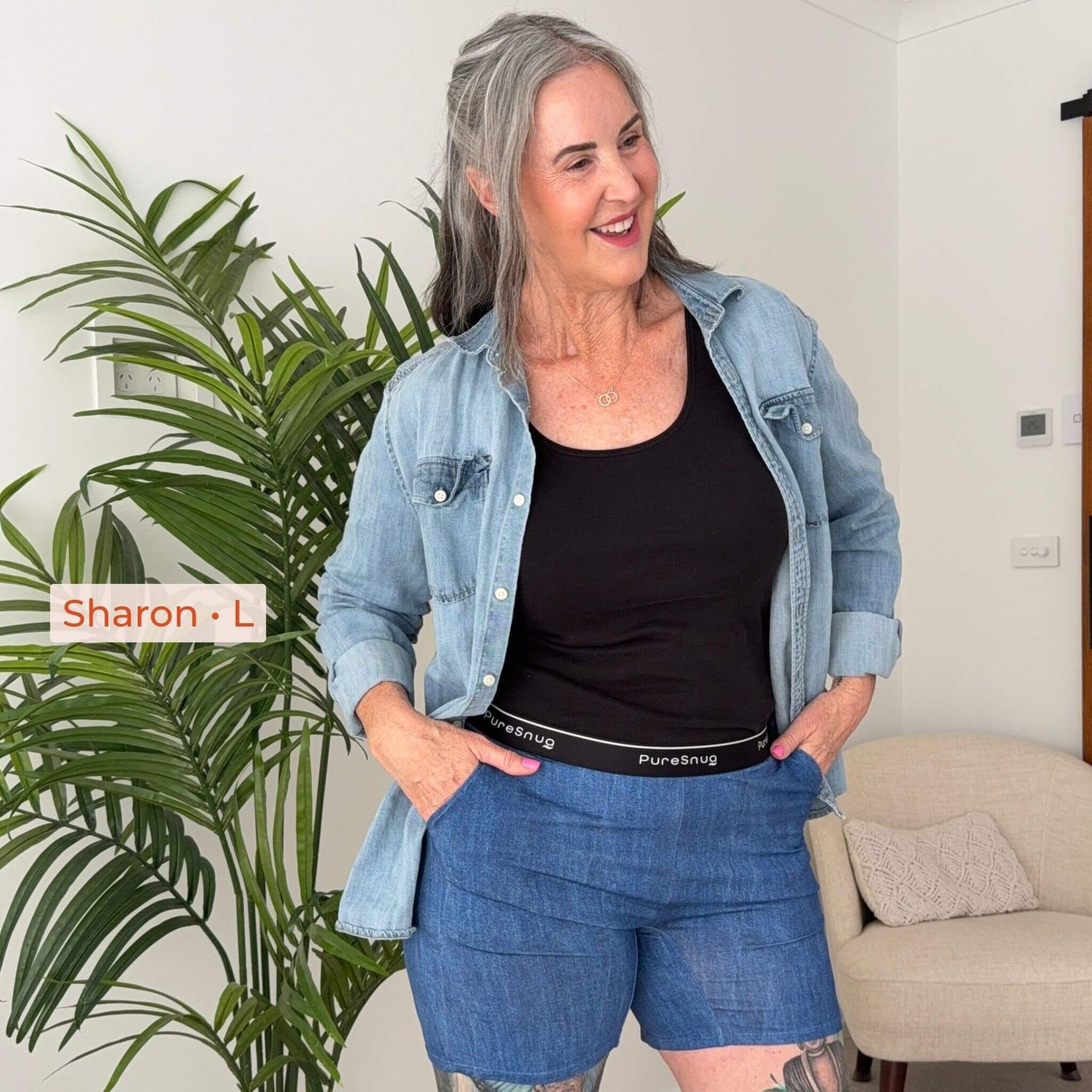 A woman wearing a black tank top, light blue denim shirt, and blue denim shorts stands indoors next to a large green plant. A text overlay reads 'Sharon - L' and the denim shorts waistband shows the brand 'Pure Snug'.