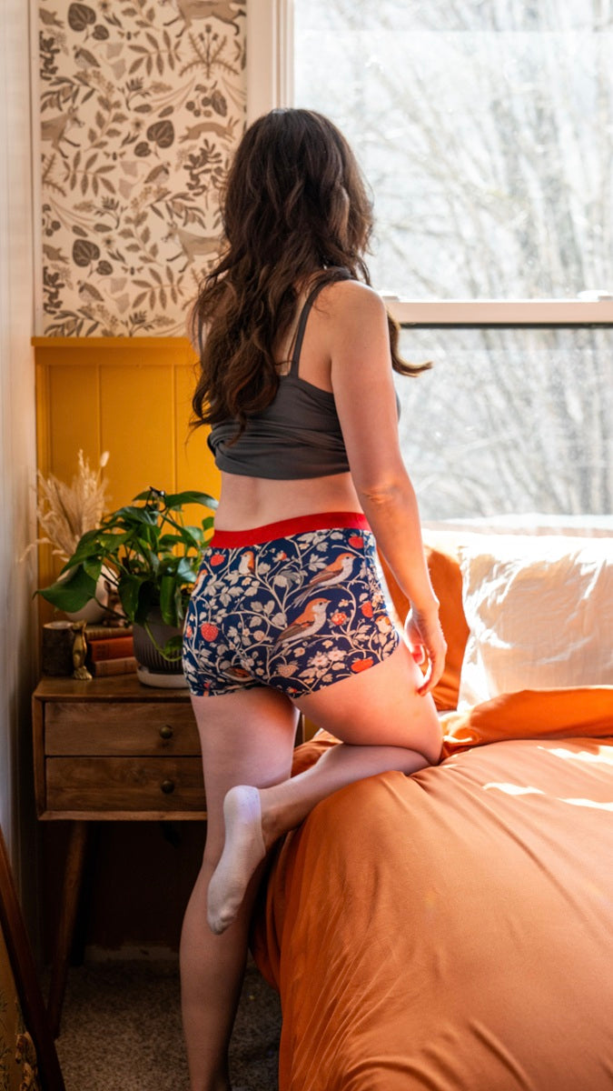 Woman in patterned Pure Snug underwear and gray tank top standing on a bed in a room with a window and decorative wall.